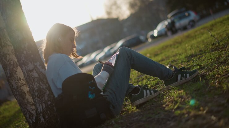 Studente si riposa all'aperto, studente si gode la lettura all'aperto al sole, persona studia tranquillamente sotto l'ombra frondosa del pomeriggio, individuo si dedica allo studio silenzioso sotto la luce intensa del sole e degli alberi