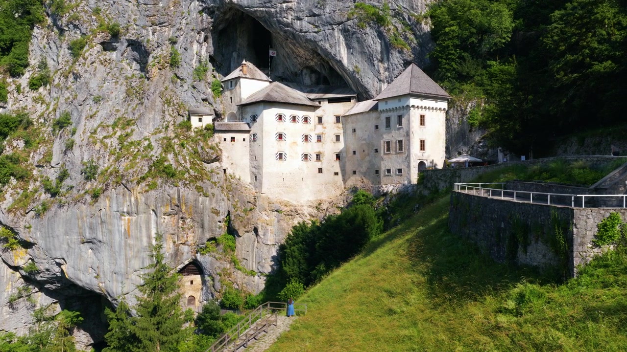 Woman Standing In Front of Predjama Castle In Inner Carniola, Slovenia. Historic Fortress Built Into A Rocky Cliff Landscape. Aerial Shot