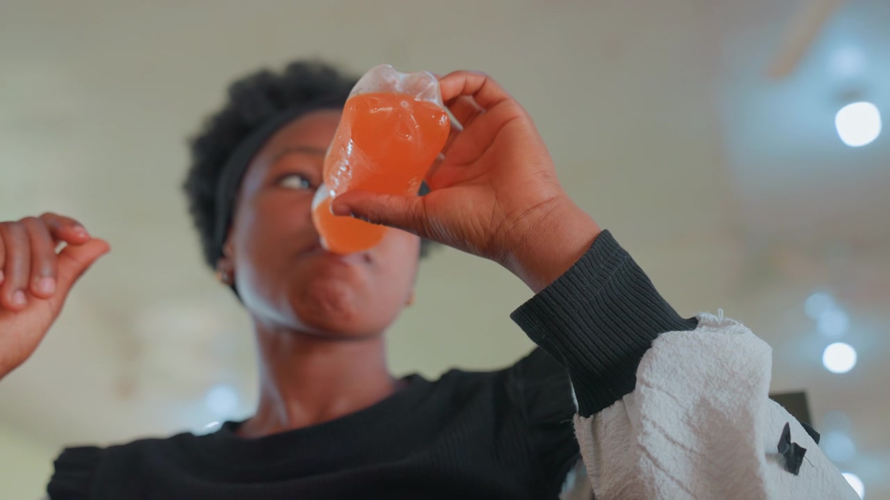 Drinking from orange bottle, refreshing sip closeup with condensation and bright highlights, candid indoor moment focused on lips and bottle, casual hydration break and vivid color contrast