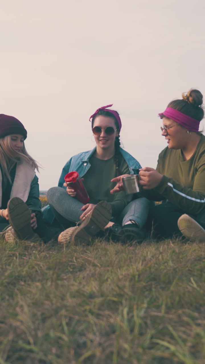 pretty girl tourists with cups of drinks sit on green grass by tent against clear sky at nice sunset time low angle shot