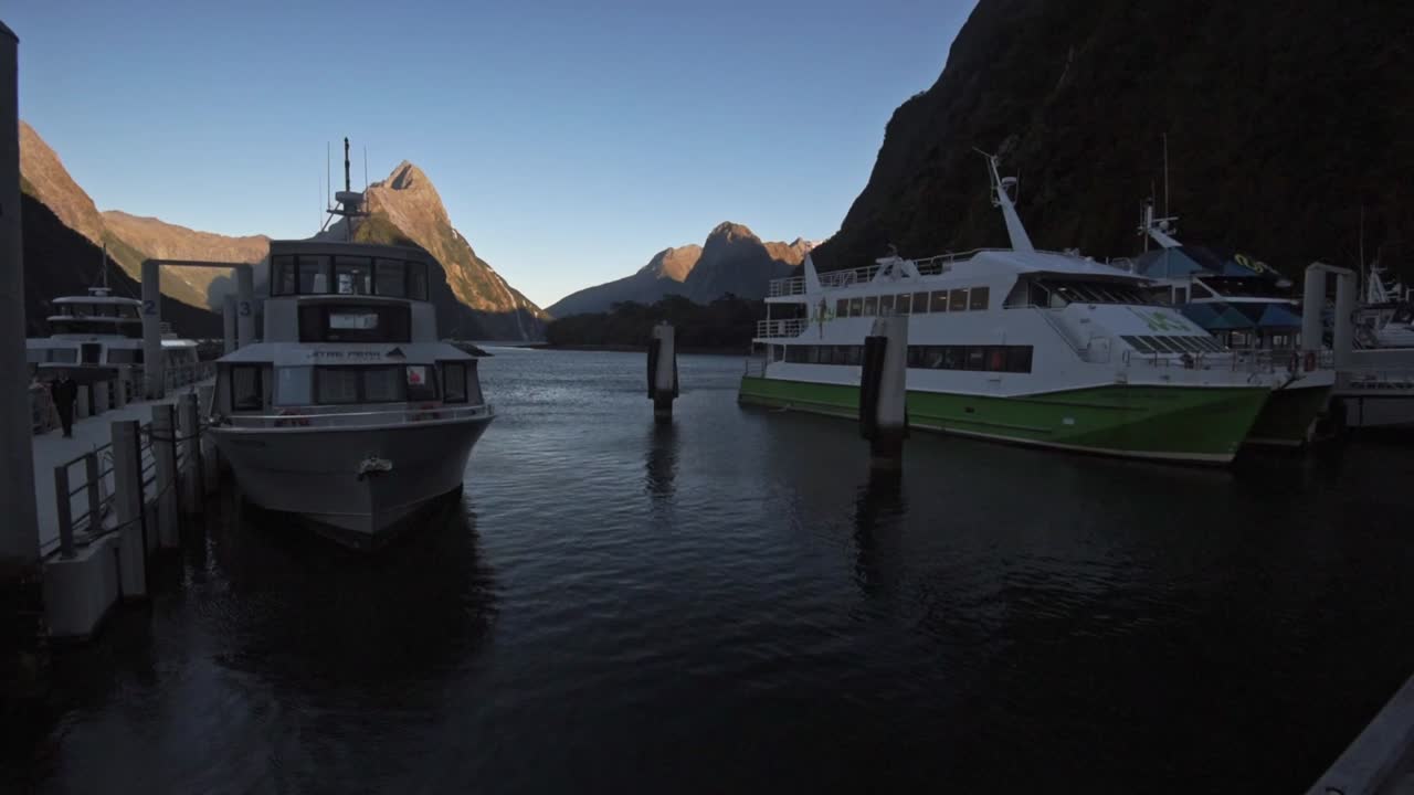Cruising ships in the harbor of Milford Sound in the foreground. Mountains and the fiord during sunrise in the background. Wide angle pan right