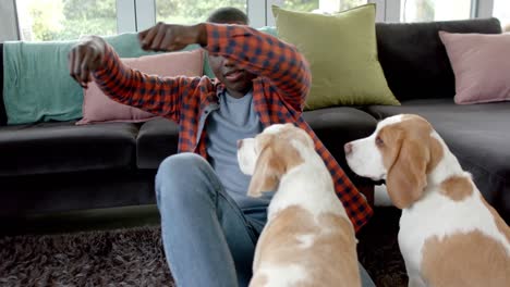 happy african american man sitting on floor at home, playing with his pet dogs, slow motion