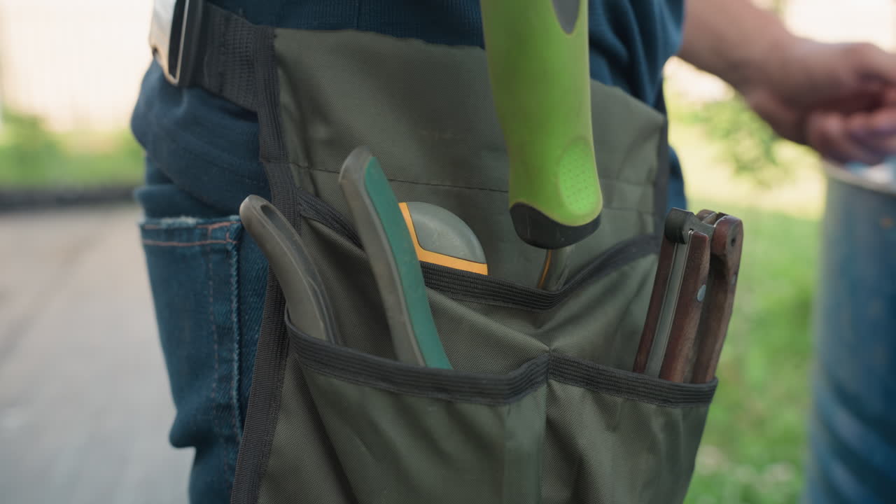 Close up of gardener organizing hand tools while trying to insert utility knife into side pocket of waist apron, surrounded by other garden tools, showing hands-on preparation before work