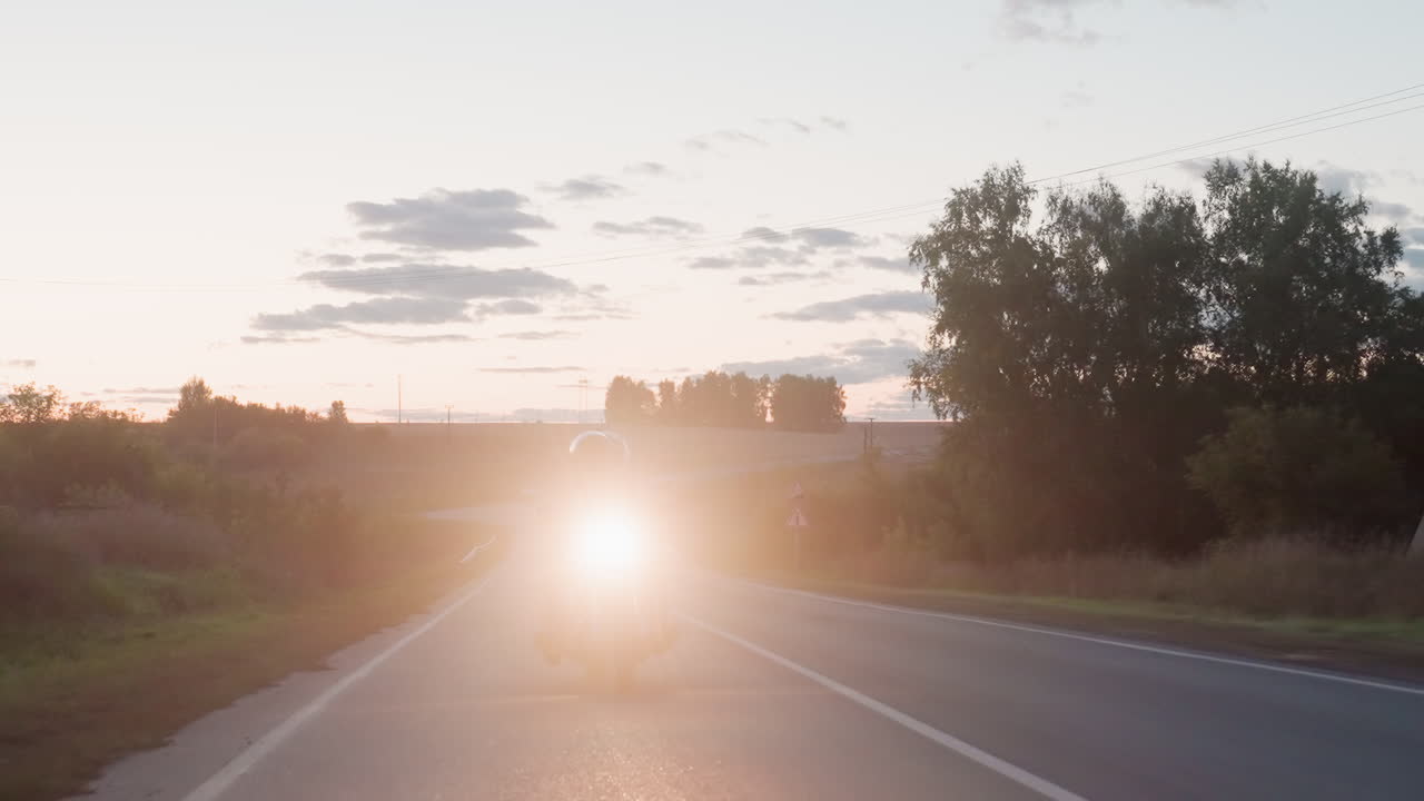 Motorcyclist wearing helmet rides powerful bike along winding countryside road at sunset while bright headlamp illuminates path ahead, tees line roadside as sky glows with warm fading evening light