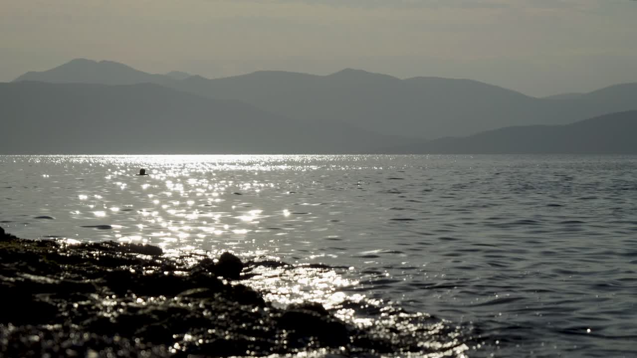 Low angle shot over sparkling ocean with mountain in distance and person swimming