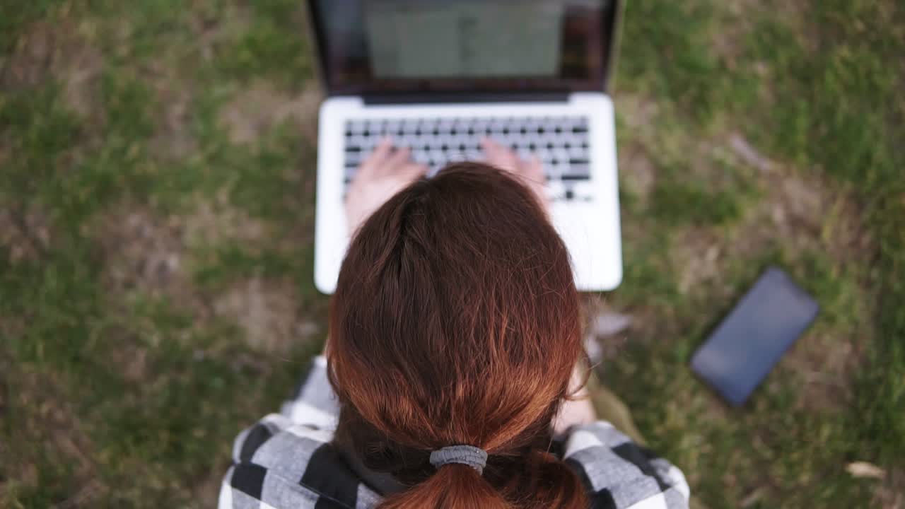 apuntando imágenes de una chica con una cola de pelo sentada en el suelo en un parque, impresiones en una computadora portátil borrosa. móvil está en la hierba cerca. disparando desde arriba hacia abajo
