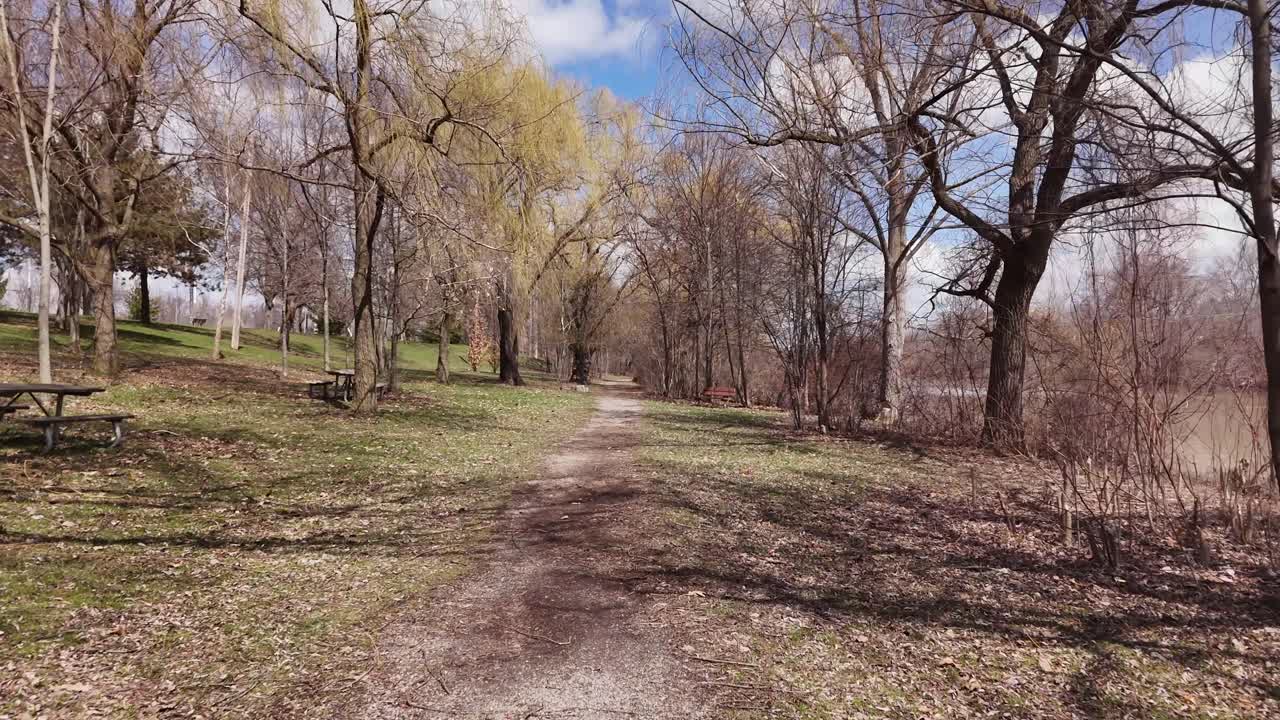 Drone che vola lungo un sentiero in un parco in primavera. Panche e tavoli da picnic fiancheggiano il sentiero con un fiume che scorre sulla destra
