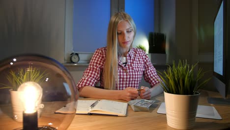 blond woman at desk looking at bitcoin. beautiful young blond female sitting at workplace with computer and open notebook at night concentrating on metal bitcoin held in hands