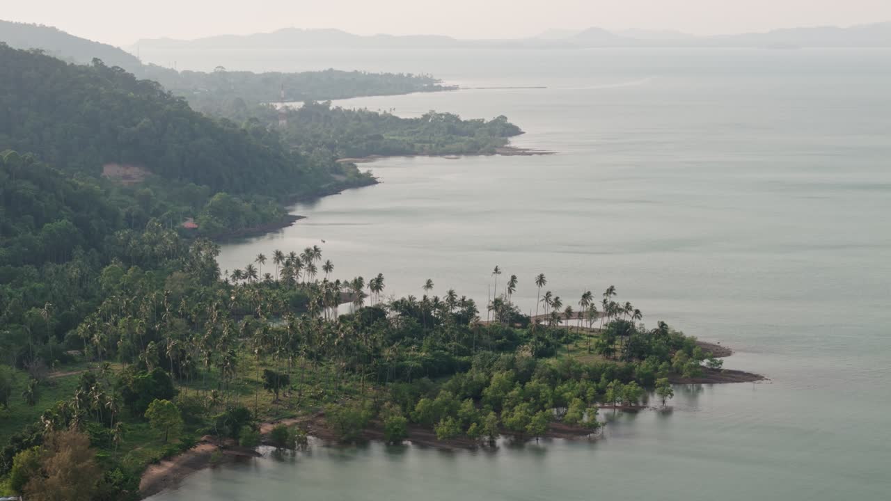 Aerial drone view descending over misty tropical coastline with palm trees and lush green hills along calm bay waters in Thailand