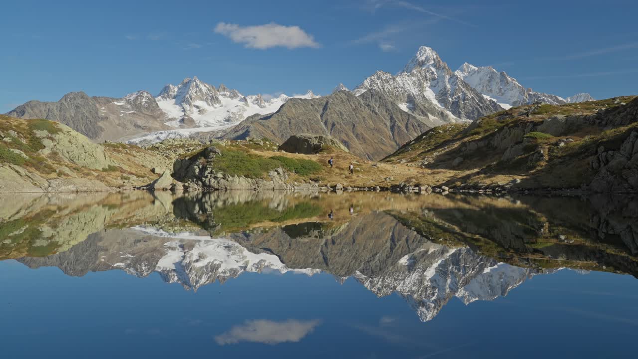 Distant hiking couple walk past lake with reflection of alpine mountains