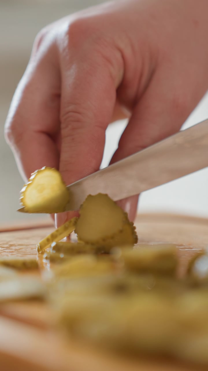 Vista ravvicinata di una mano che impila sottili fette di olive verdi su un caldo tagliere di legno con un piccolo coltello, mostrando un'attenta preparazione in cucina, una consistenza lucida e un delicato movimento verso il basso.