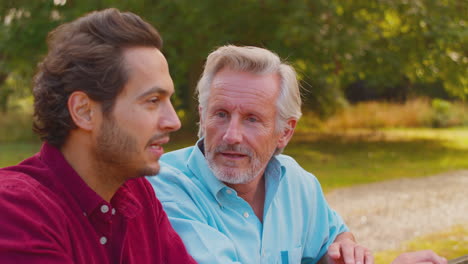 Mature-Father-With-Mixed-Race-Adult-Son-Leaning-On-Fence-Walking-In-Countryside