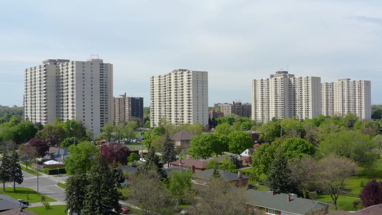 Aerial drone footage capturing multiple apartment buildings overlooking a peaceful suburban neighborhood with mature trees, detached homes, and landscaped residential streets under clear daylight.
