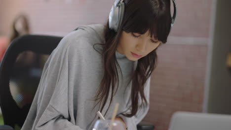 young asian business woman writing notes enjoying study listening to music wearing headphones female student drinking juice in office workplace close up