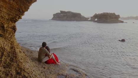 Couple-Sitting-On-Beach-Enjoying-Beautiful-Seascape,-Rocks-Formation-In-Water