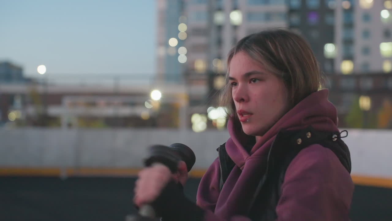 Wellness minded woman boxing dumbbells punching air on black asphalt court next to white barrier topped with chain link fence under urban dusk sky wearing maroon hoodie white sneakers
