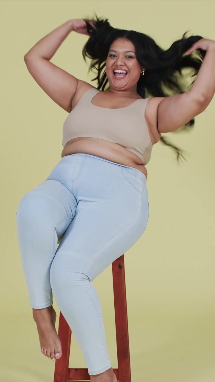 Video verticale: Con le mani sui fianchi, una donna in canotta e jeans si muove in studio su uno sgabello per un servizio fotografico.