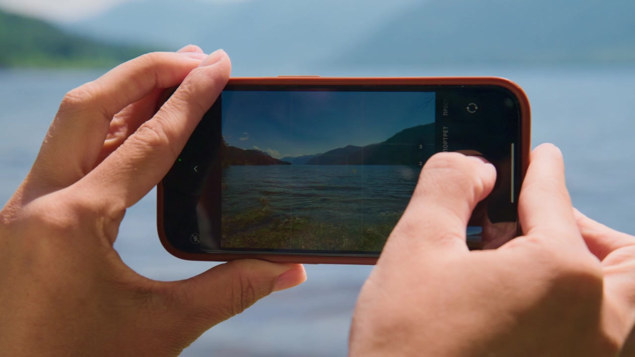 Mãos seguram smartphone, capturando vista panorâmica do lago. Dedos ajustam o foco da câmera na tela. A imagem mostra água calma, montanhas distantes. Ideal para temas de viagem, natureza ou tecnologia.