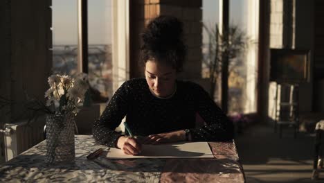 Joven-Artista-Femenina-Sofisticada-Dibujando-Con-Un-Bolígrafo,-Sentada-Junto-A-La-Mesa-En-Un-Estudio-De-Arte-Con-Amplias-Ventanas-Panorámicas-En-El-Fondo.-Inspiración,-Plena-Concentración.