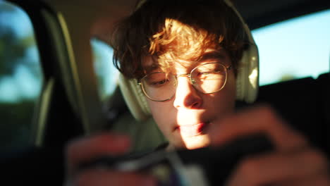 teenage passenger deeply immersed in mobile gaming, wearing headphones while traveling inside moving vehicle during golden hour light