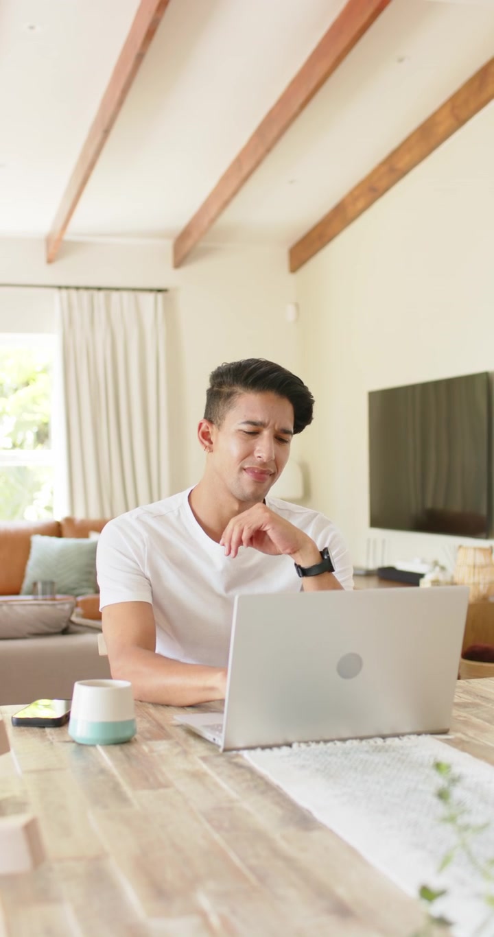 縦型動画：ダイニングテーブルで、困惑した様子のラップトップメッセージを読み、腕時計を確認し、返信を入力している男性。