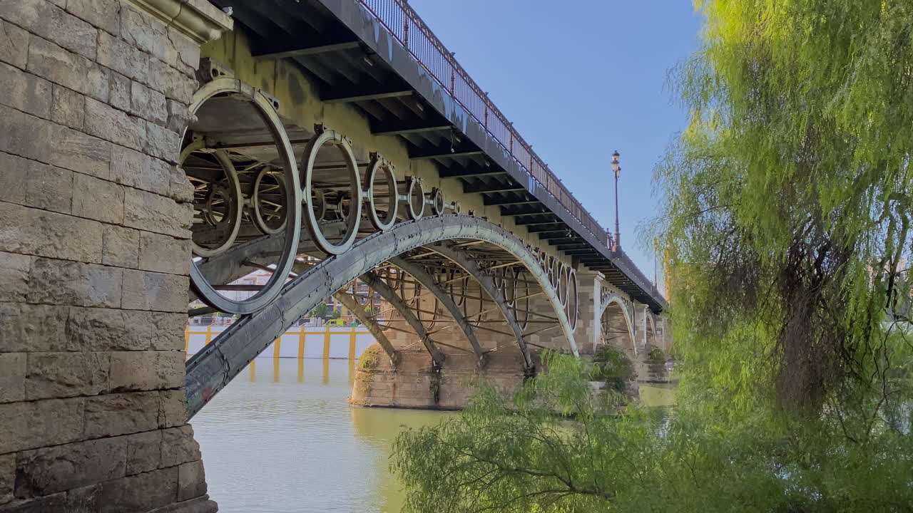 Triana-silta vehreän kasvillisuuden ja kirkkaan nurmikon läpi nähtynä, rauhallinen jokinäkymä aurinkoisessa Sevillassa