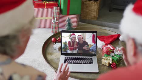 Pareja-Caucásica-Mayor-Usando-Una-Computadora-Portátil-Para-Una-Videollamada-Navideña-Con-Una-Pareja-Feliz-En-La-Pantalla