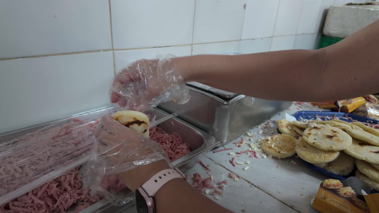 close-up food service worker's gloved hands preparing Venezuelan arepas in a commercial setting. Hands are seen scooping shredded ham (jamón) and cheese from metal containers to stuff arepas