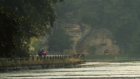 Gente-En-Bicicleta-En-La-Orilla-Del-Lago-Harasov-Con-Una-Montaña-Rocosa-En-La-Distancia-En-Kokorin,-República-Checa