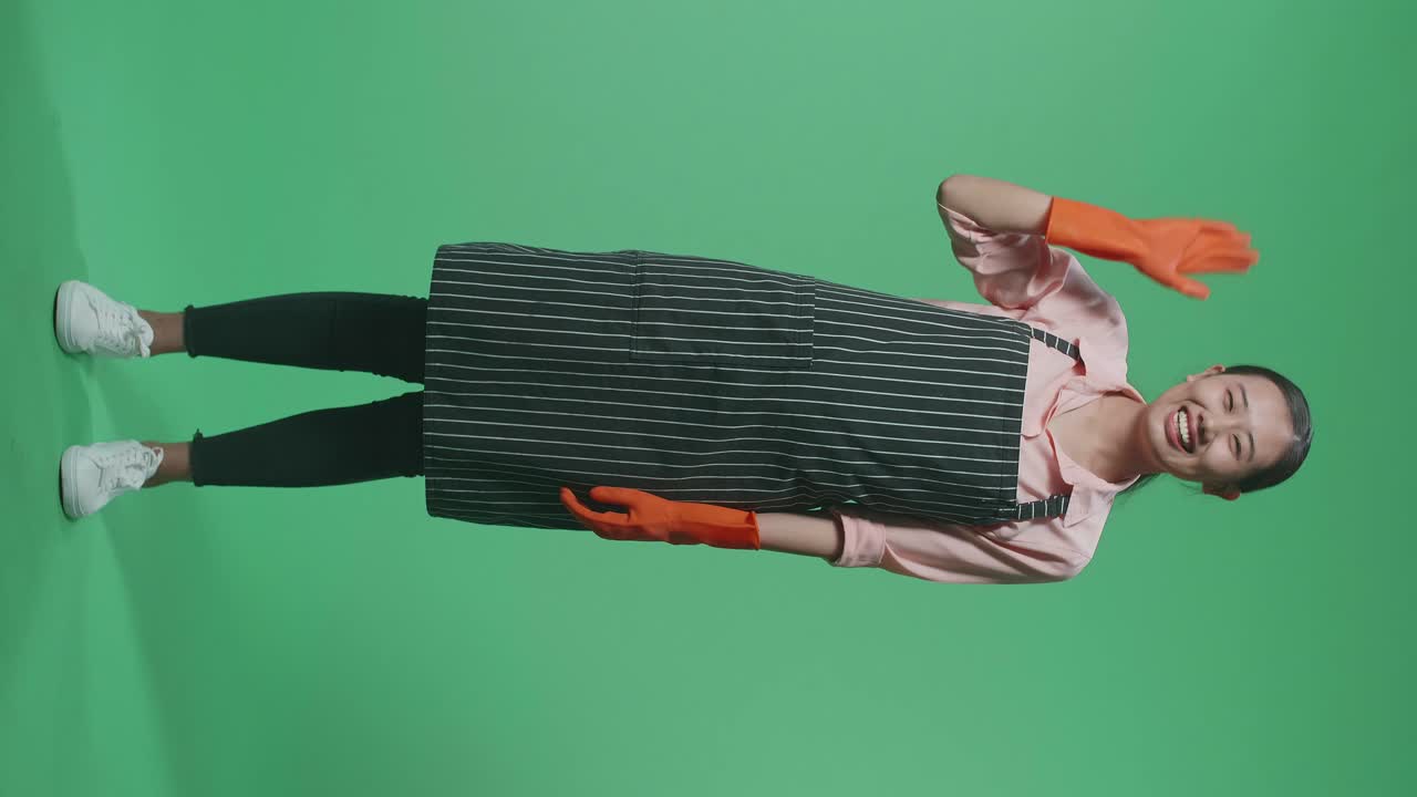 초록색 스크린 스튜디오에 서 있는 동안 손을 흔들고 미소 짓는 아프론을 입은 아시아 여성 집주인의 몸이 가득 차 있습니다.