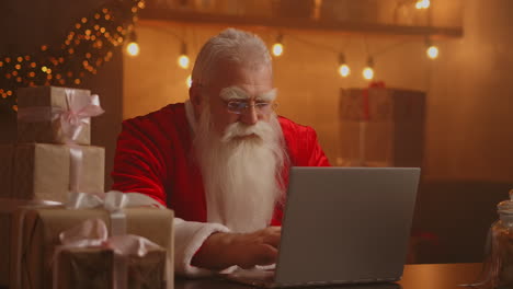 Santa-Claus-holding-gift-box-typing-keyboard-kid-talking-to-child-greeting-on-Merry-Christmas-Happy-New-Year-in-virtual-online-chat-on-laptop-sitting-at-home-table-late-with-present-on-xmas-eve.-High-quality-4k-footage