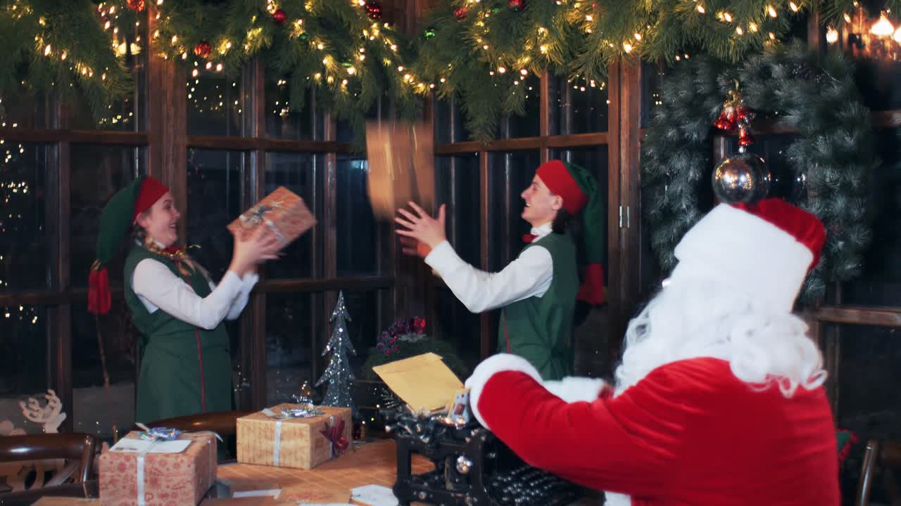 Santa Claus sitting at a table in a decorated room typing on an old typewriter. Elves play with gift boxes. New Year's residence Santa Claus