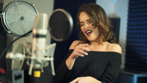 expressive woman with curly hair is singing and dancing in front of a microphone in an audio recording studio on a blue background. close-up