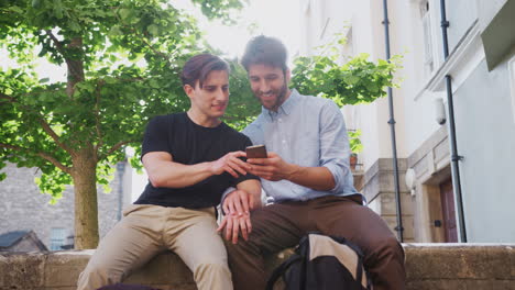 Una-Pareja-Gay-Masculina-Sentada-Al-Aire-Libre-En-La-Pared-Del-Edificio-Mirando-El-Teléfono-Móvil