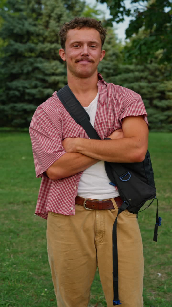 Young Gen Z man standing outside with arms crossed, smiling confidently and casually. Friendly modern lifestyle, authentic youth portrait, natural light, vertical format