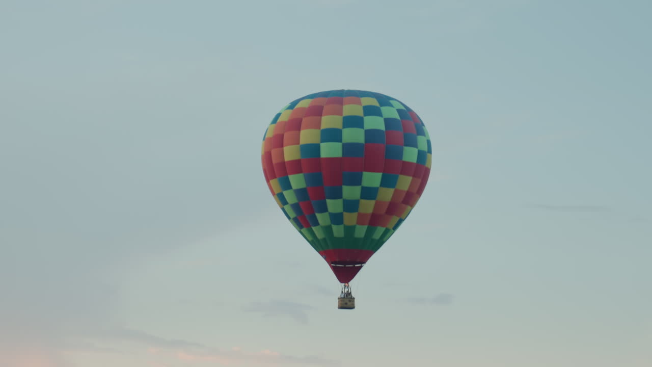 distant view of multicolored hot air balloon drifting gently over vast green fields filmed from high vantage point under soft pastel sky at golden hour