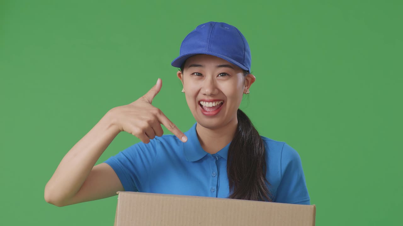 Close Up Of Asian Female Courier In Blue Uniform Smiling And Pointing To A Carton In Hand While Delivering It On Green Screen Background In The Studio