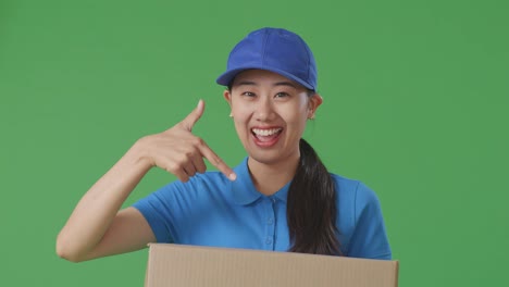 푸른 유니폼을 입은 아시아 여성 택배기사가 미소 짓고 손에 박스를 가리키며 스튜디오의 녹색 화면 배경에 전달합니다.