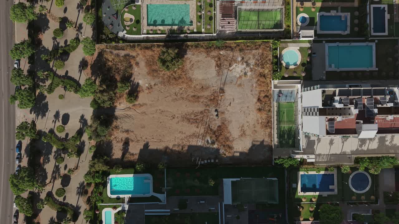Top-down aerial view of an urban block featuring a vacant construction plot surrounded by residential buildings with pools, trees, and organized landscaping