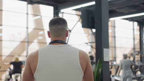 shoulder view of guy walking toward gym equipment station with blurred exercisers in background under bright led lights and industrial interior featuring steel beams and large windows