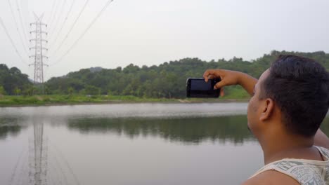 man boy in tank top taking video panoramic shot of transmission tower power grid power line by the lake over the mountain reflection in water like a mirror in mobile