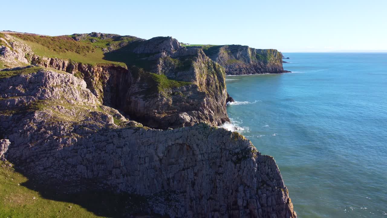 Descending Aerial View of Various Different Coastal Cliff Formations with Beautiful Calm Blue Sea and Steep Mountains. Gower Peninsula in South Wales, UK