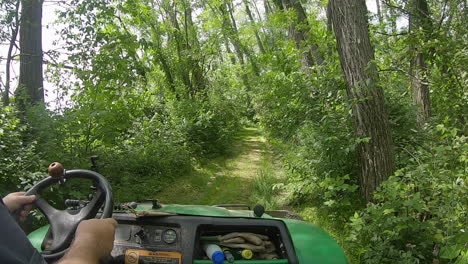 view of hands and steering wheel of a man driving a utv on a grassy trail through lush woods along a fence row