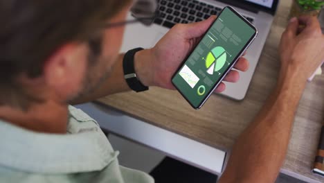 hombre caucásico sentado en un escritorio usando un teléfono inteligente con estadísticas en la pantalla