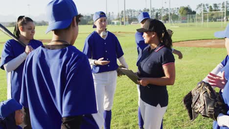 diverse group of female baseball players listening to female coach with clipboard talking on pitch