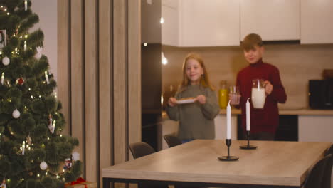 ein blonder junge und seine blonde schwester stellen ein leeres glas, einen krug milch und einen teller voller kekse auf einen leeren tisch mit zwei kerzen, dann verlassen sie raum 2