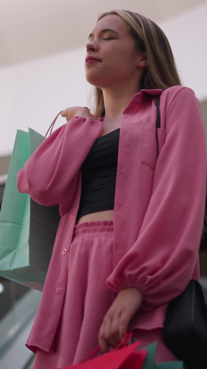 dama con una camiseta negra bajo un traje rosa, sosteniendo una bolsa de compras verde menta detrás de ella y otra bolsa en su mano derecha mientras está en una escalera mecánica moviéndose hacia arriba, en el interior de un centro comercial