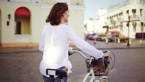 Back-view-of-a-woman-in-a-white-shirt-and-blue-jeans-walking-in-the-city-street-holding-her-bicycle-handlebar-with-a-basket