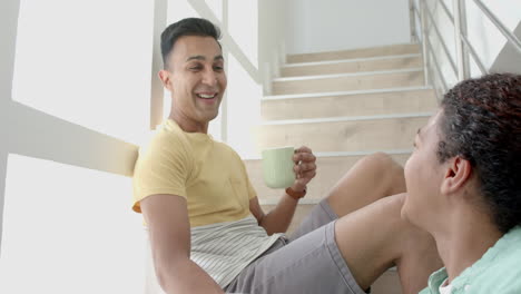 Happy-diverse-gay-male-couple-sitting-on-sunny-staircase-having-coffee-and-talking,-slow-motion