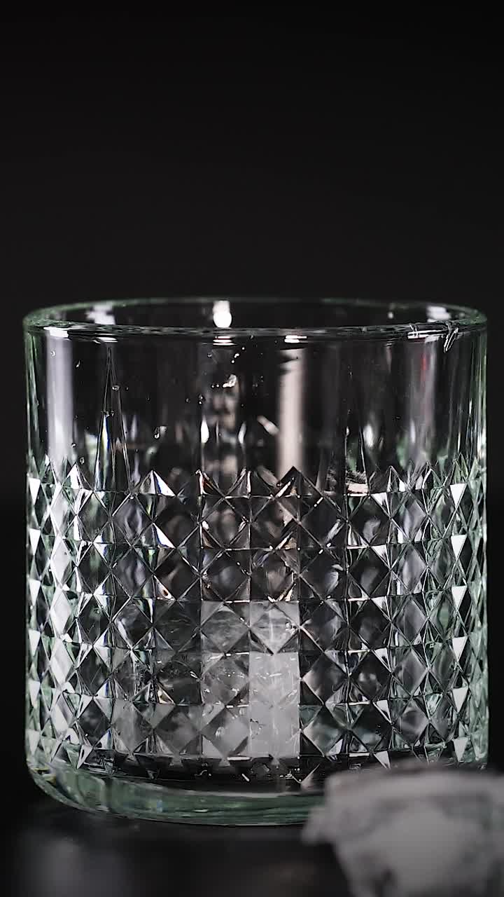Optagelse i slowmotion af isterninger, der falder ned i et tekstureret krystalglas mod en mørk baggrund.